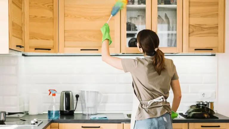 Woman dusting upper wooden cabinets while organizing kitchen for deep cleaning.