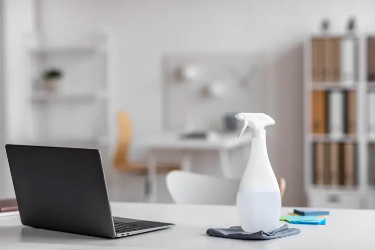 White spray bottle and microfiber cloth placed on a white desk next to a laptop in a bright home office.