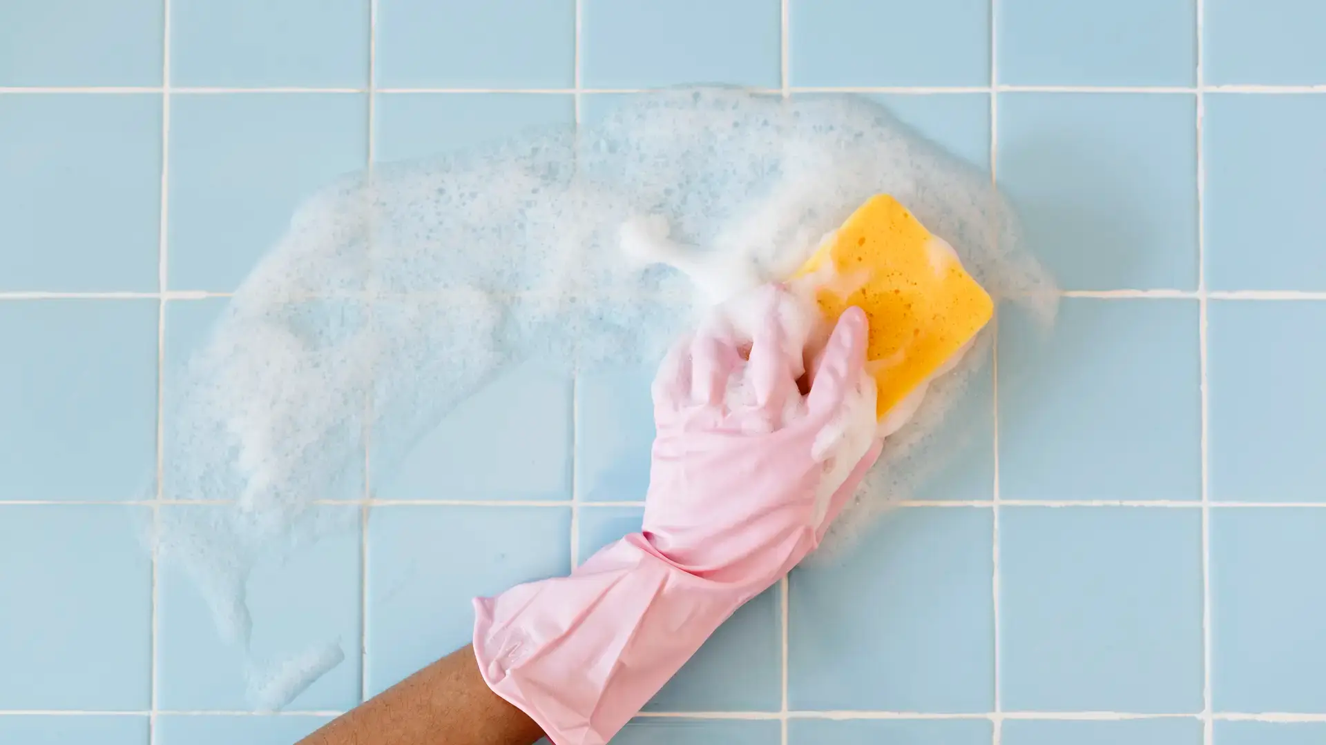 Hand in glove scrubbing soapy tile wall with sponge